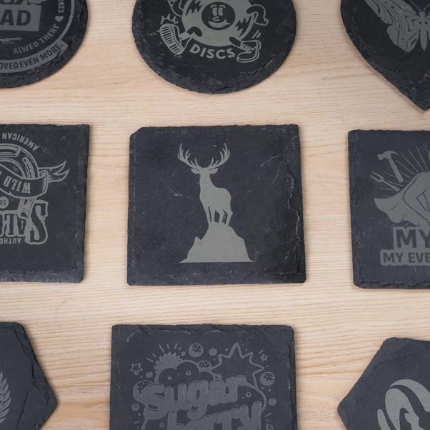 An assortment of custom laser-engraved dark slate coasters in round, square, and hexagonal shapes, featuring detailed designs including a stag silhouette, logos, and stylised graphics on a light wooden surface.