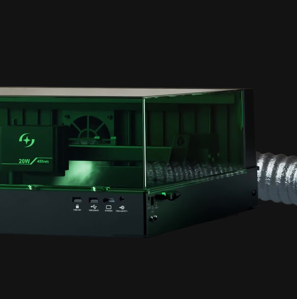 The enclosed AtomStack Fusion laser engraver featuring a green protective shield with smoke accumulating inside, showing a flexible exhaust hose connected to the back for ventilation.