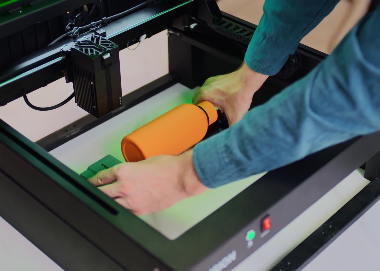 A user placing a bright orange water bottle onto a rotary roller attachment inside an AtomStack Fusion laser engraver, which is elevated on its riser base.