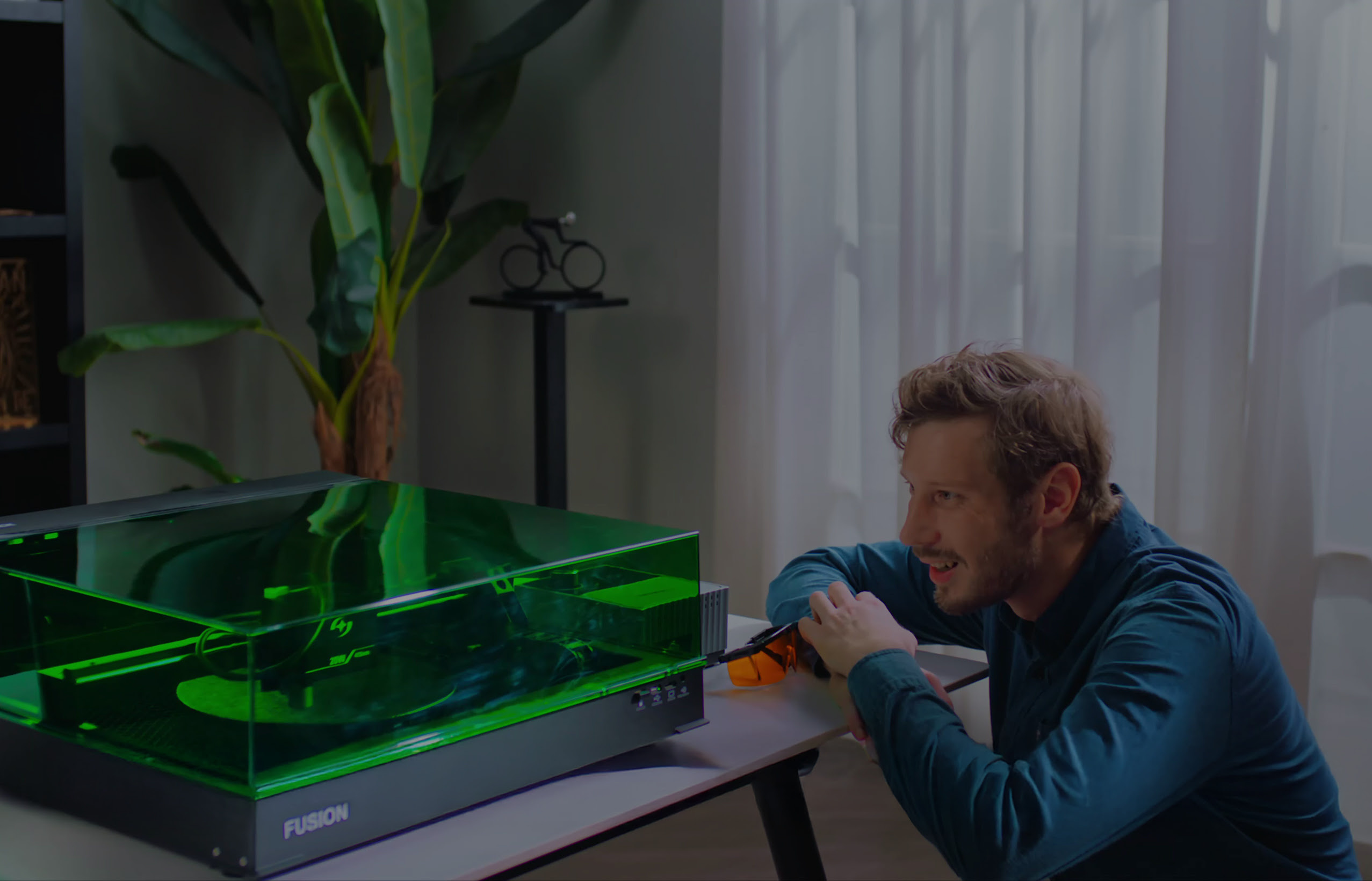 A man looking closely at an enclosed AtomStack Fusion laser cutter operating under its protective green safety cover on a desk in a home setting.
