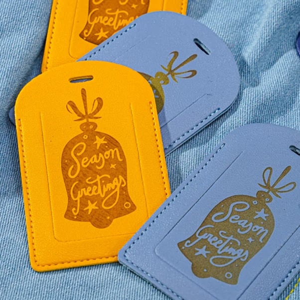 Four leather cardholders, in yellow and periwinkle blue, are fanned out on a denim background. They are engraved with a "Seasons Greetings" bell motif.