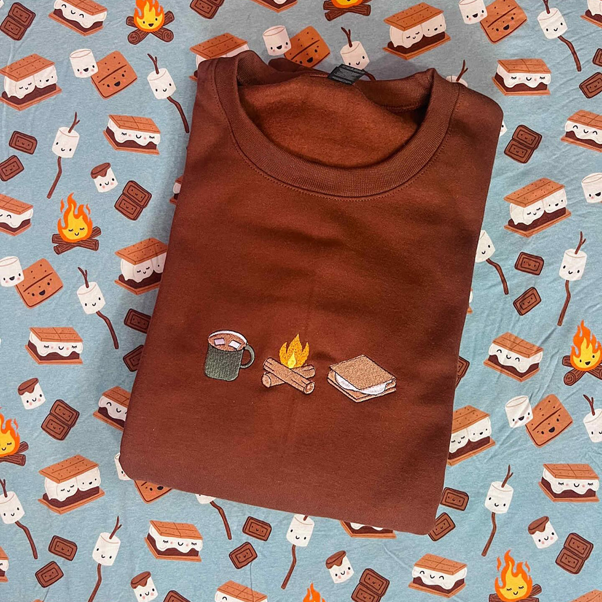 Brown shirt with an embroidered design of a cup of hot chocolate with marshmellows, a campfire and a s'more