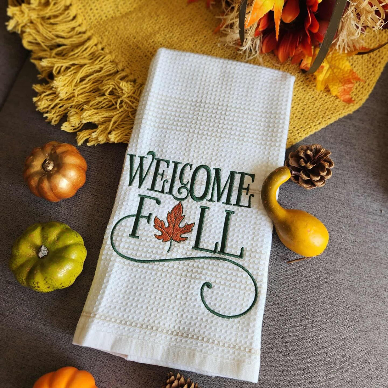 Kitchen towel with an embroidered design of the text Welcome Fall in ornate green lettering.
