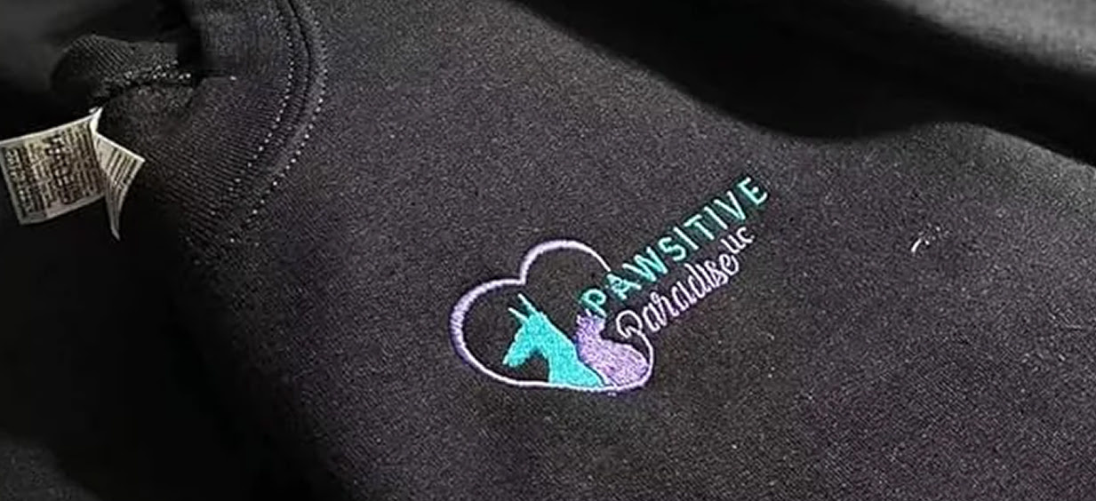 Close-up of a black shirt with an embroidered design of a cat and dog in a heart shape and the text: Pawsitive Paradise.