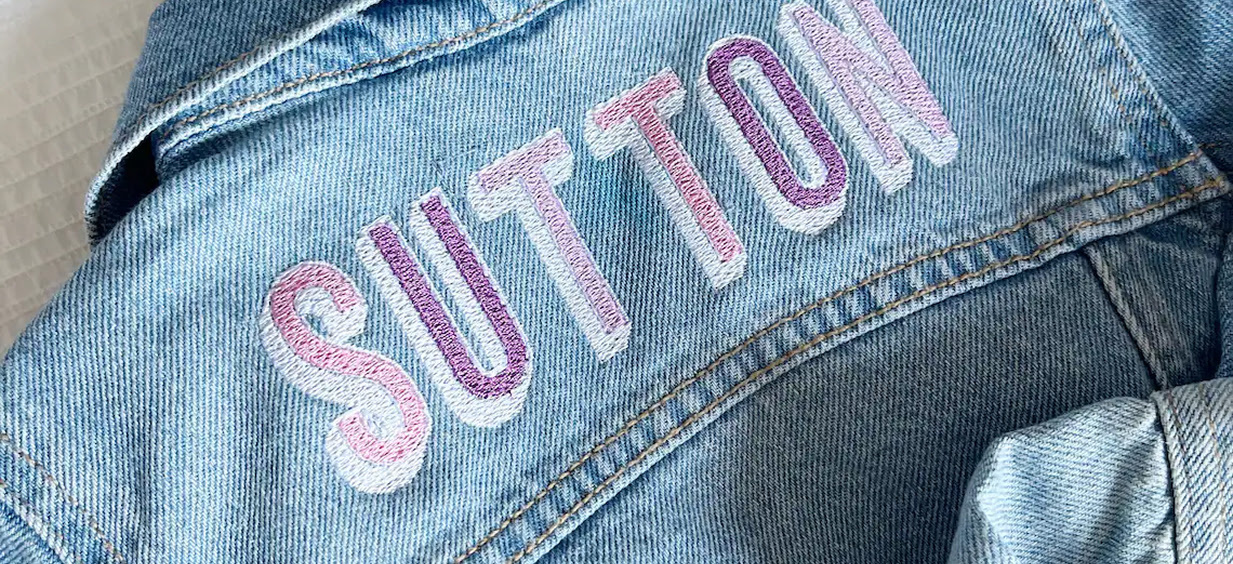 Custom light-wash denim jacket featuring the name SUTTON embroidered across the back yoke in a block letter font with a purple and pink gradient.
