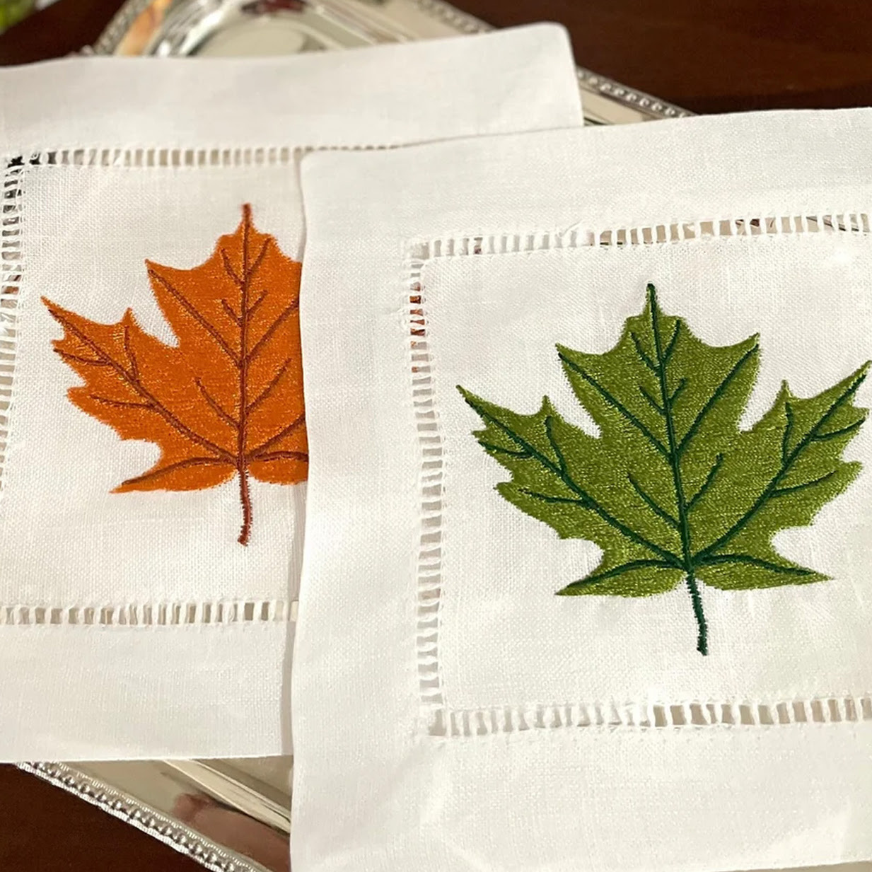 Two white linen cocktail napkins with ladder-stitch borders, each featuring a detailed embroidered maple leaf: one in autumn orange and one in green.