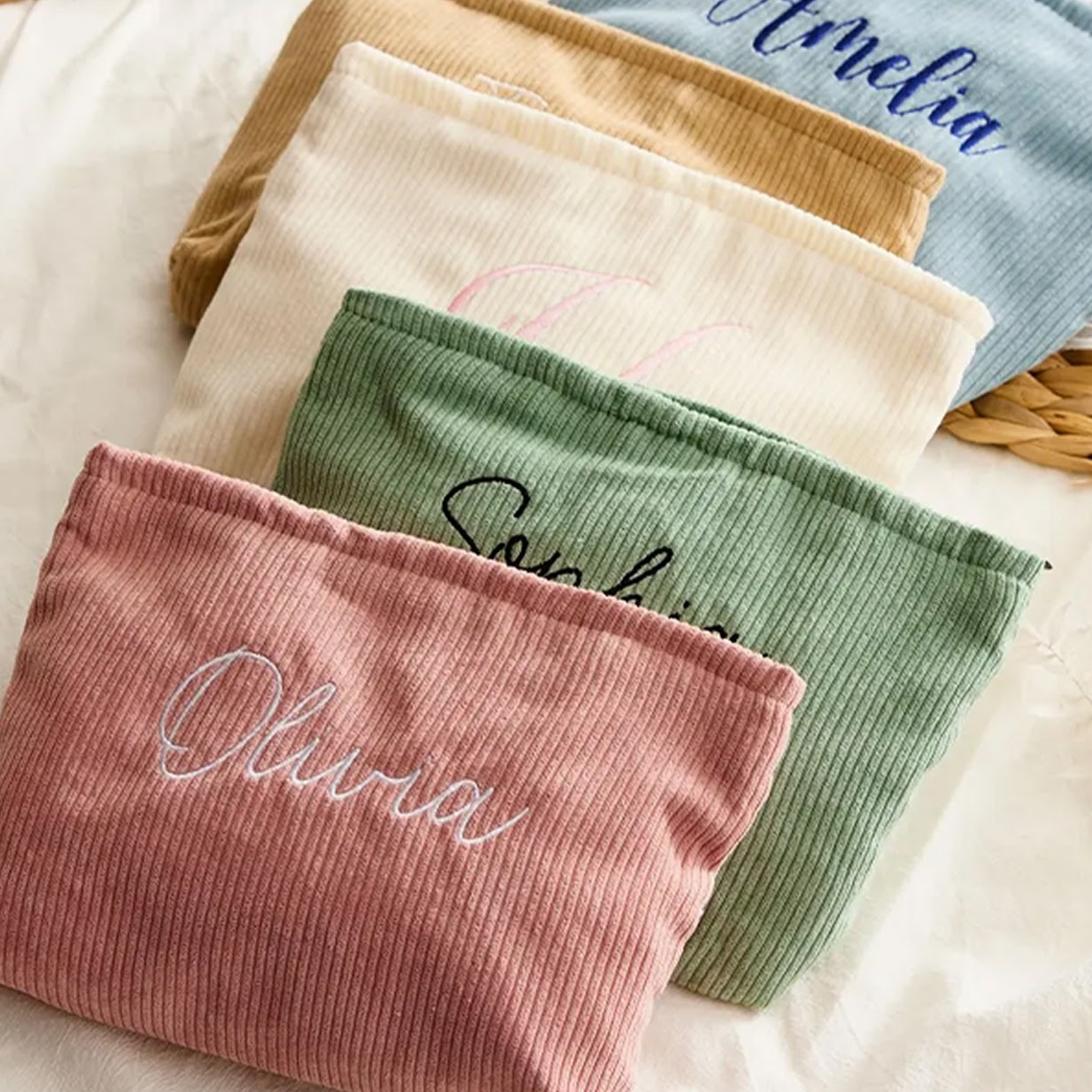 A collection of personalised corduroy pouches in pastel colors, featuring custom embroidered names like Olivia and Amelia in script font.