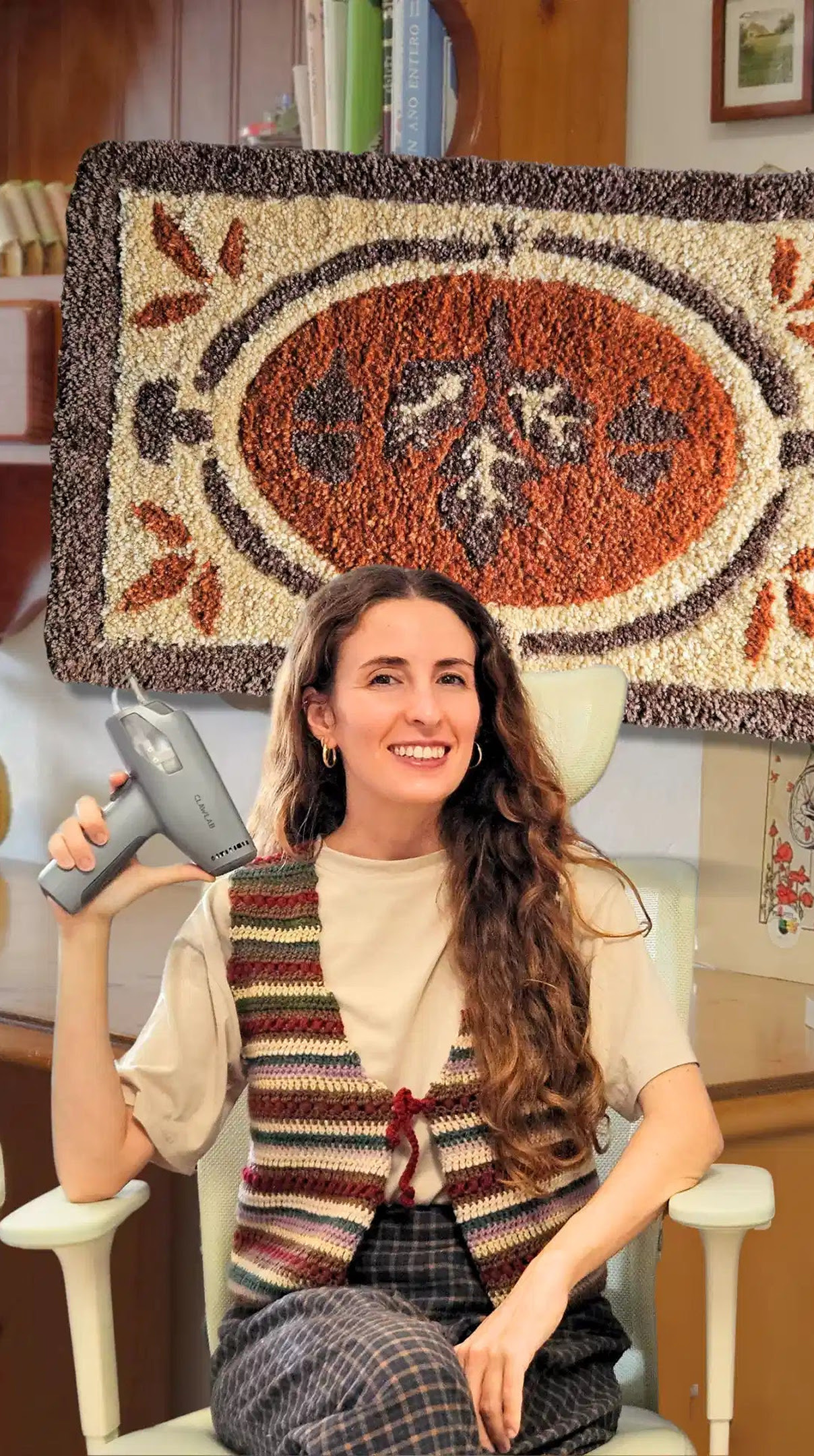 Woman holding a tufting gun in front of a tufted rug with a floral design in brown, cream and grey yarn.