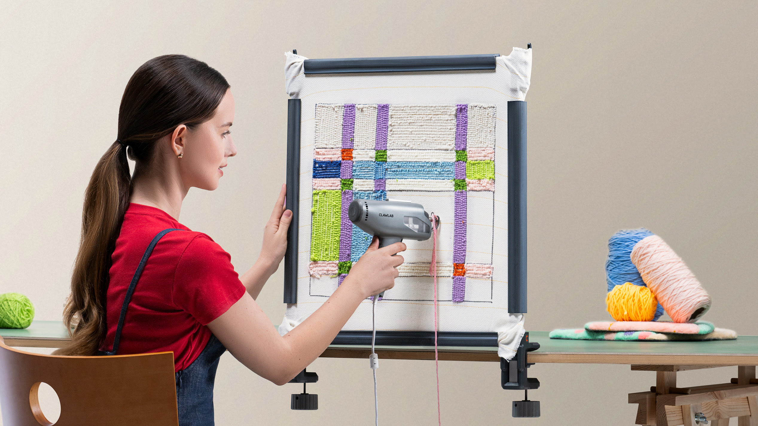 Woman sitting at a desk using a Clawlab tufting gun on a table-clamped tufting frame with colourful yarn spools nearby