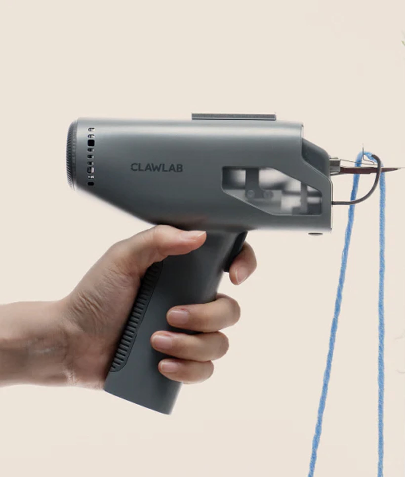 Hand holding a grey Clawlab electric tufting gun threaded with blue yarn against a plain background.