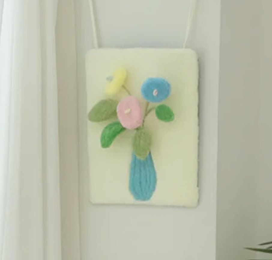 Pastel tufted floral wall hanging featuring 3D yarn flowers in a blue vase hanging on a white wall.