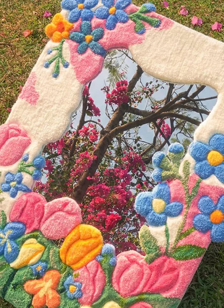 Handmade tufted floral mirror frame decorated with colourful 3D yarn flowers reflecting pink blossoms outdoors.
