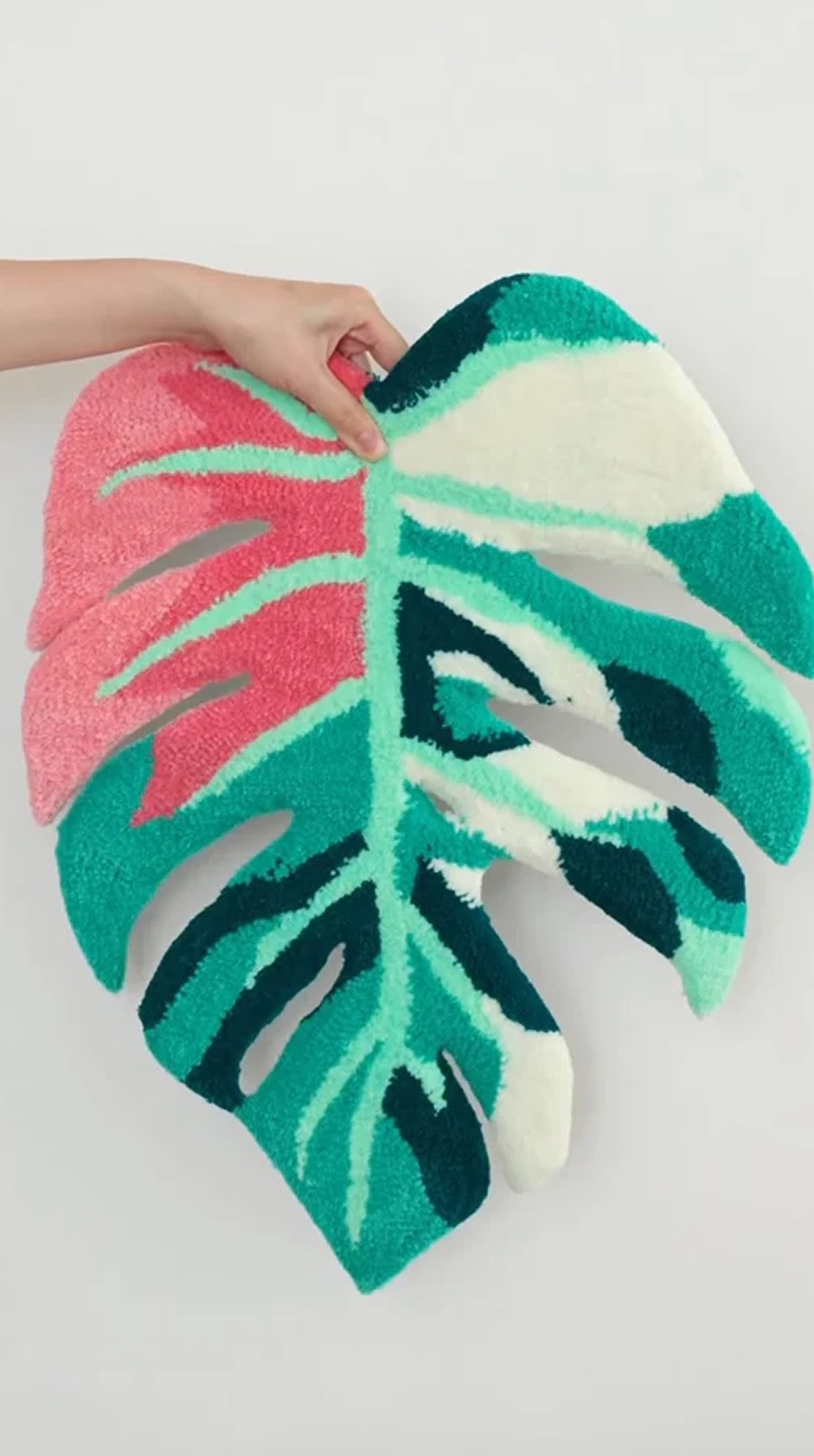 Person holding a tufted monstera leaf rug with pink, teal, green, and cream yarn patterns.